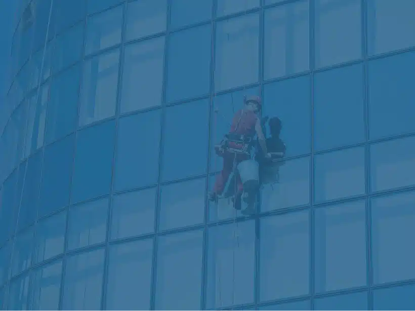Man in a harness with a red cleaning suit and hardhat, cleaning office windows high up in the air.
