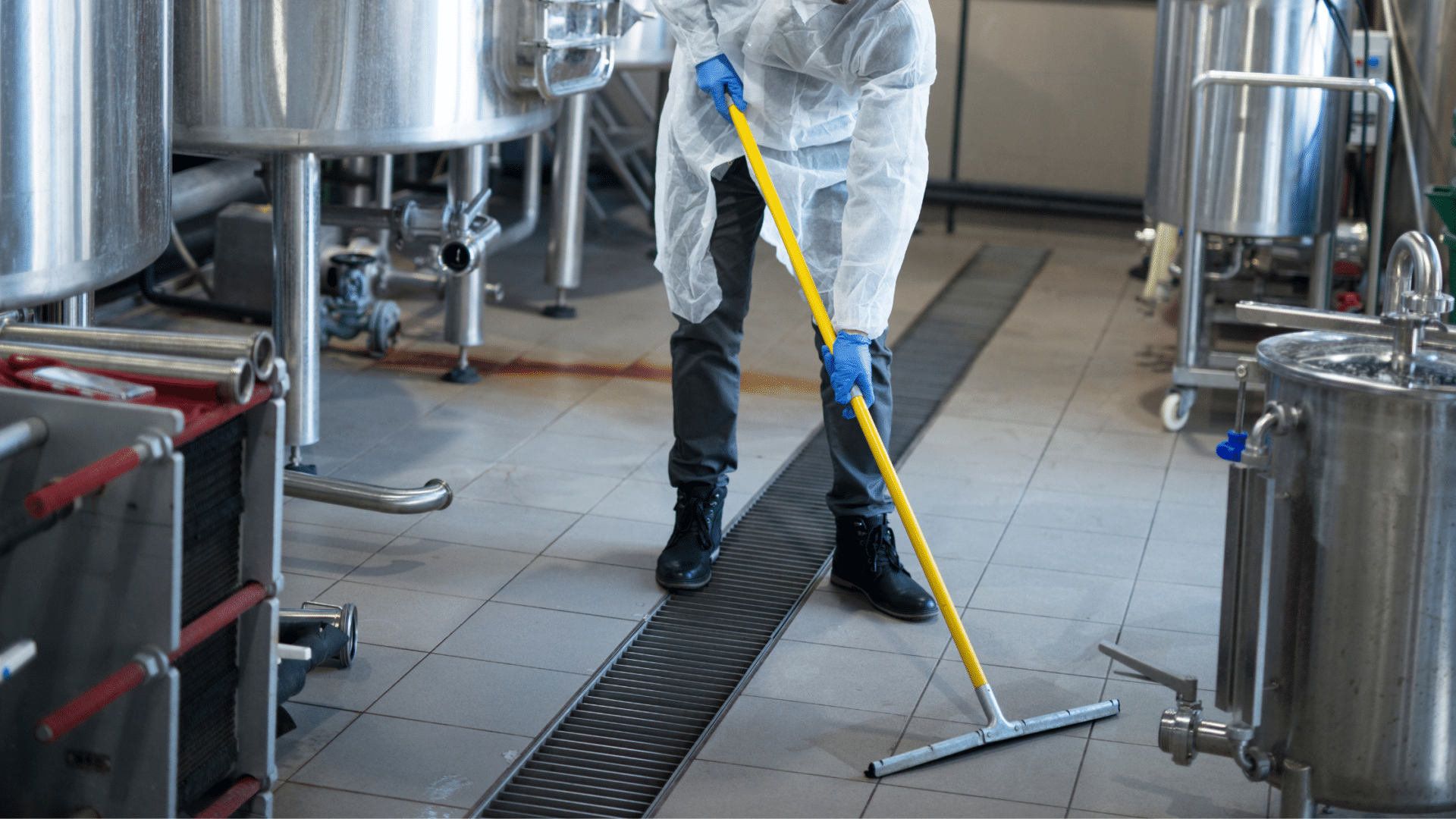 Front image of a person with a squeegee, sanitising a tiled floor with food processing equipment.