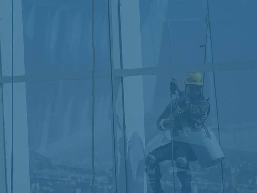 Mirror image of a man in a harness, hard hat and jacket with a bucket cleaning windows in a high-access environment.