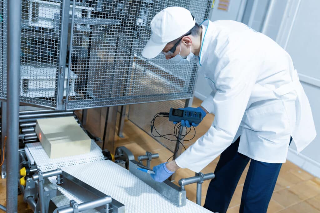 A food safety technician in a white lab coat, cap, mask, and blue gloves inspecting a conveyor belt.