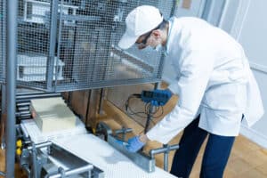 A food safety technician in a white lab coat, cap, mask, and blue gloves inspecting a conveyor belt.